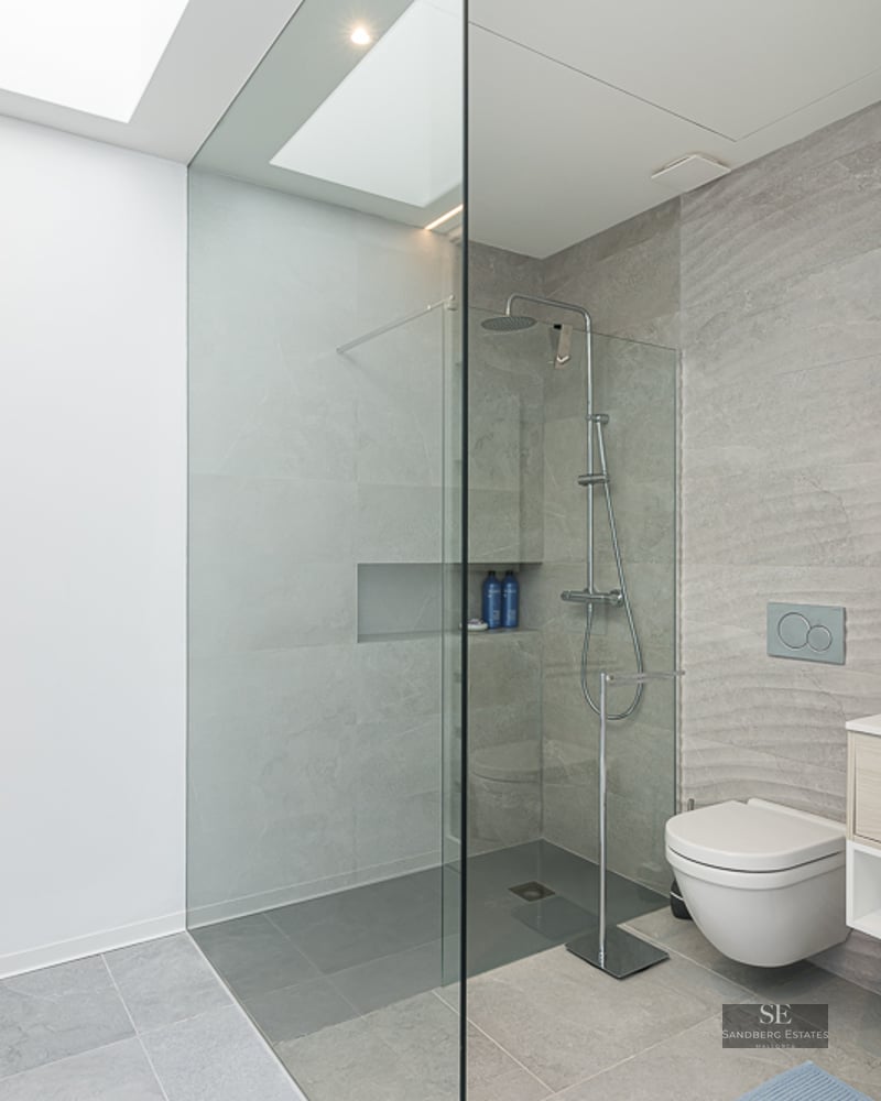 Minimalist bathroom featuring grey stone tiles, a glass walk-in shower, wall-hung toilet, and a bright skylight.