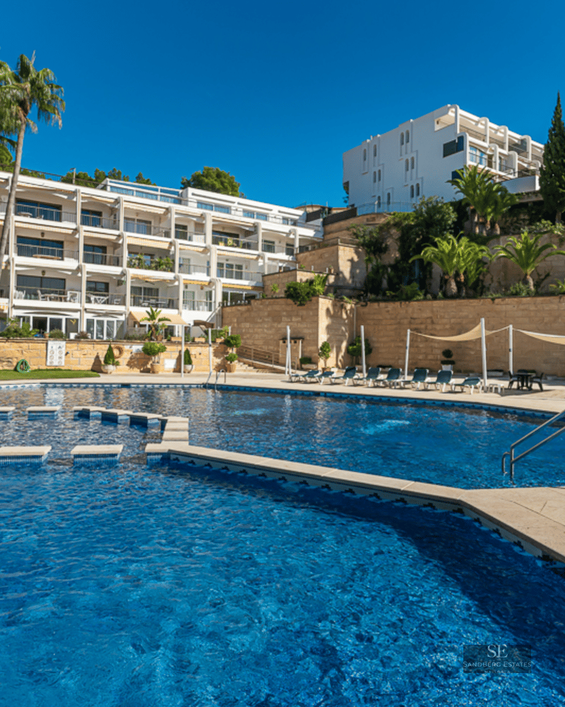 Large blue swimming pool surrounded by a stone deck, palm trees, and a white apartment building under a clear sky.