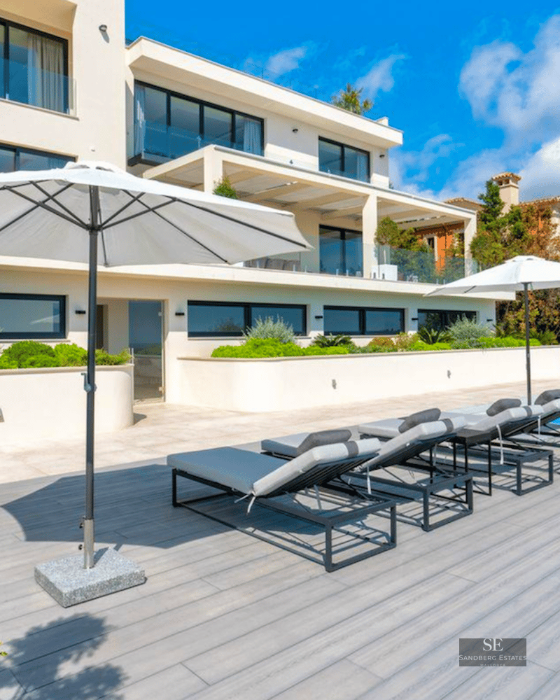 Modern villa exterior featuring a turquoise swimming pool, grey sun loungers with white umbrellas, and lush landscaping.