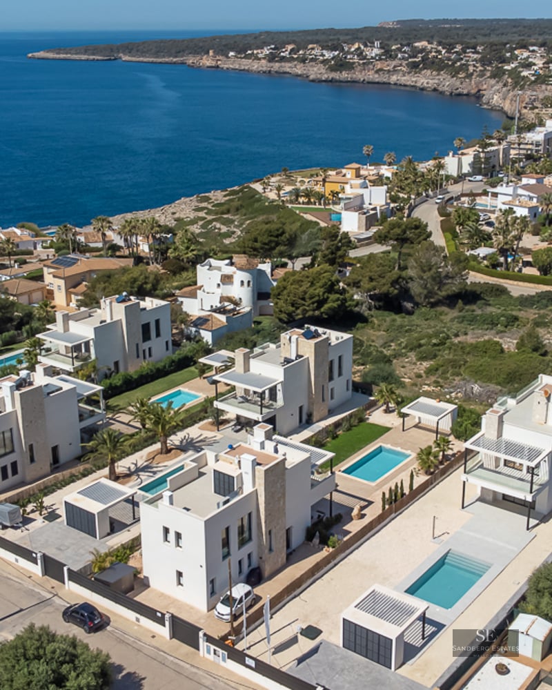Drone view of modern white villas with private pools on a rocky coastline next to the blue sea.