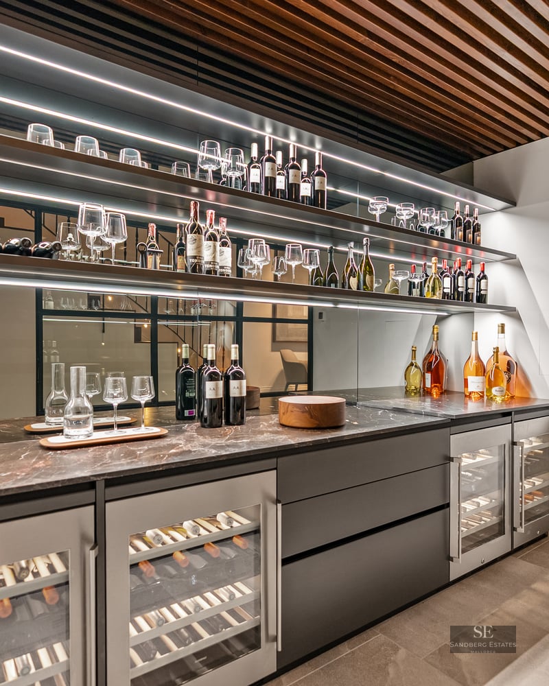 Modern wine cellar with illuminated shelves, wine coolers, marble counters, and a wooden slatted ceiling.