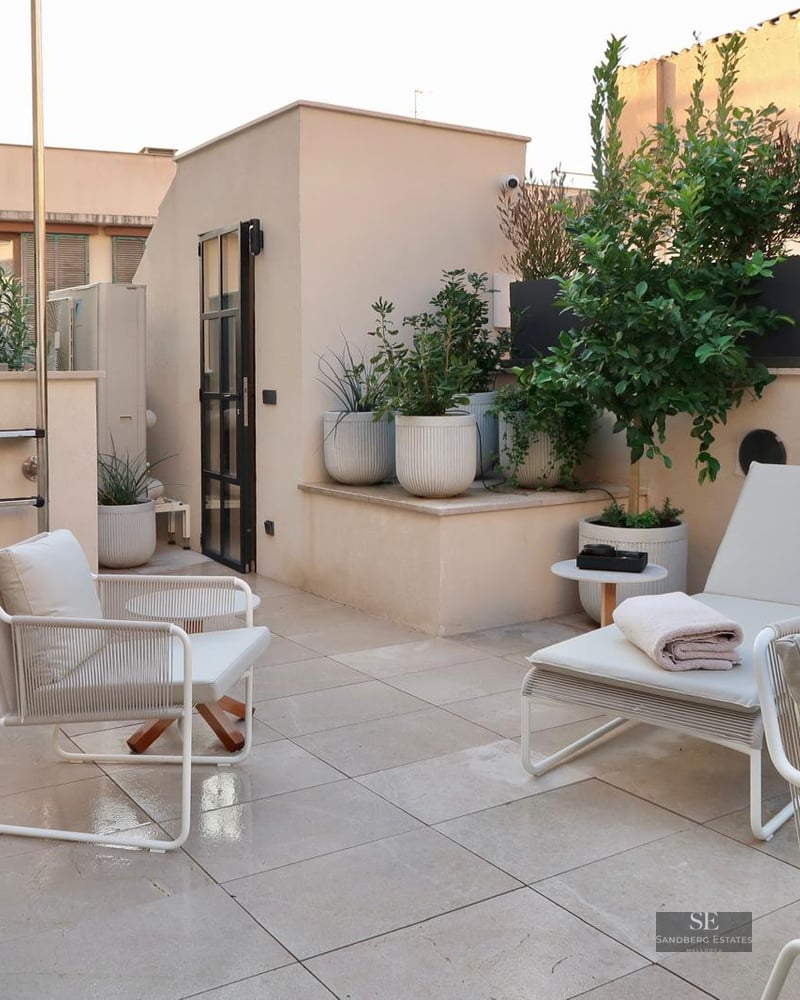 A chic rooftop terrace with white designer armchairs, a sun lounger, potted green plants, and light-colored floor tiles.