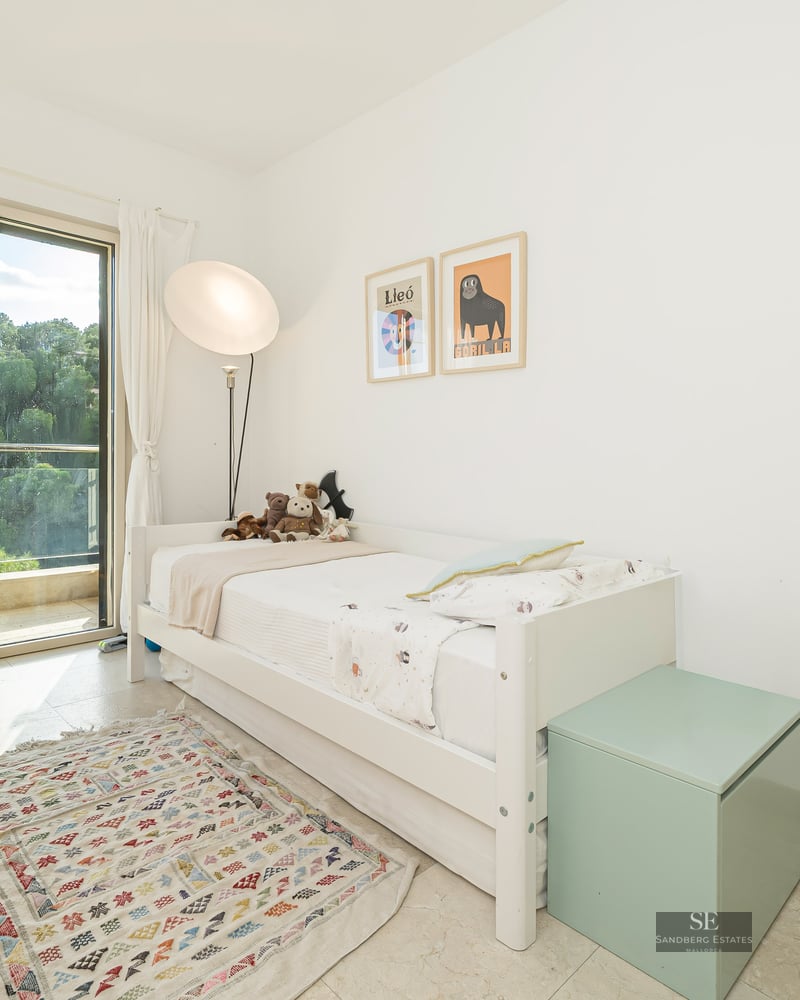 White bedroom featuring a single bed, desk, sliding glass door to a balcony, and a geometric rug.