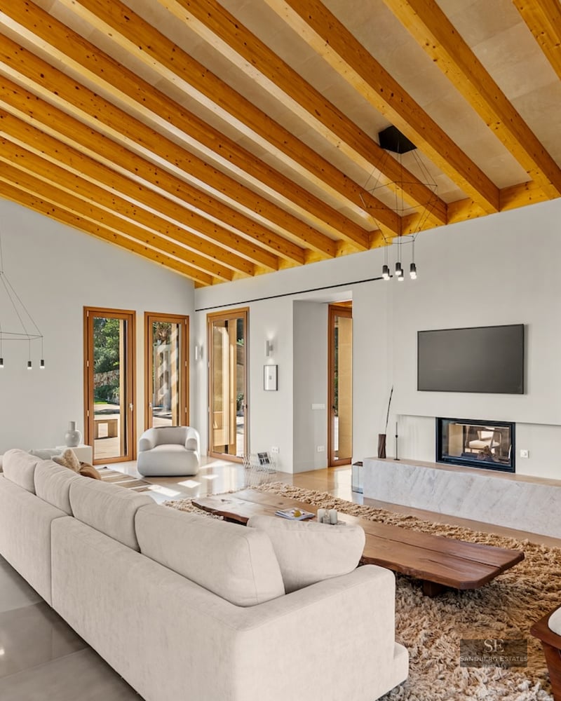 Spacious modern living room featuring high ceilings with wood beams, a large sofa, fireplace, and garden views.