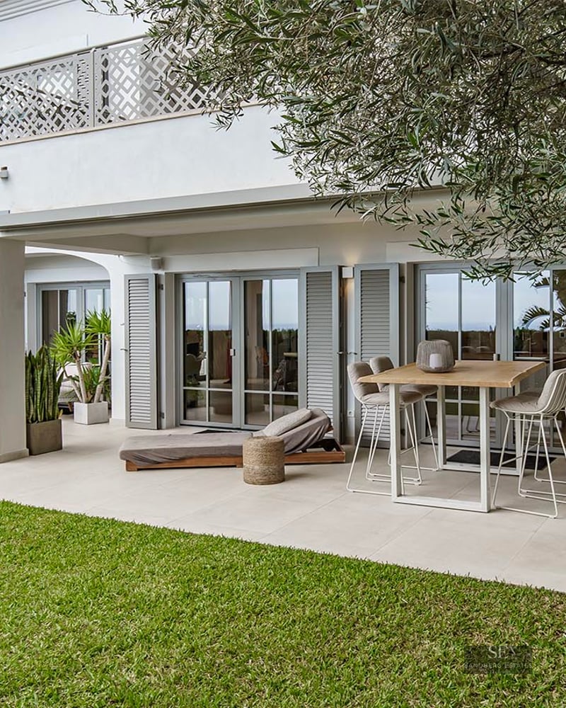 White Mediterranean villa with a covered patio, high dining table, sun lounger, green lawn, and palm trees.