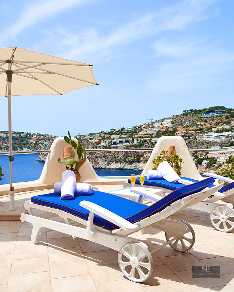 Two blue sun loungers and a white umbrella on a stone terrace overlooking the Mediterranean sea and coastline.