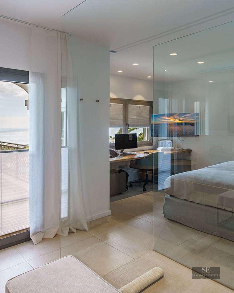 Modern bedroom with floor-to-ceiling glass wall, integrated office area, and balcony overlooking the sea.