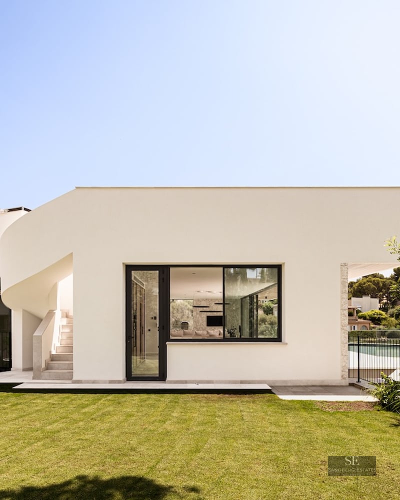 A bright white modern villa with a curved staircase and large windows overlooking a flat green lawn under a blue sky.