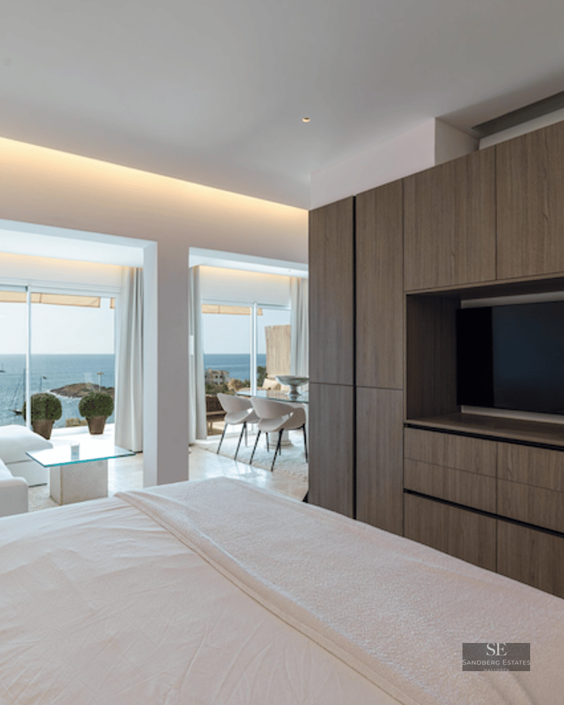 Master bedroom featuring a large wooden wardrobe with TV, white bed, and floor-to-ceiling windows overlooking the ocean.