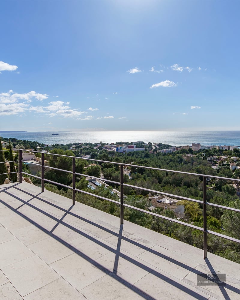 Large white stone terrace with a black metal railing overlooking a panoramic view of the Mediterranean Sea and pine trees.
