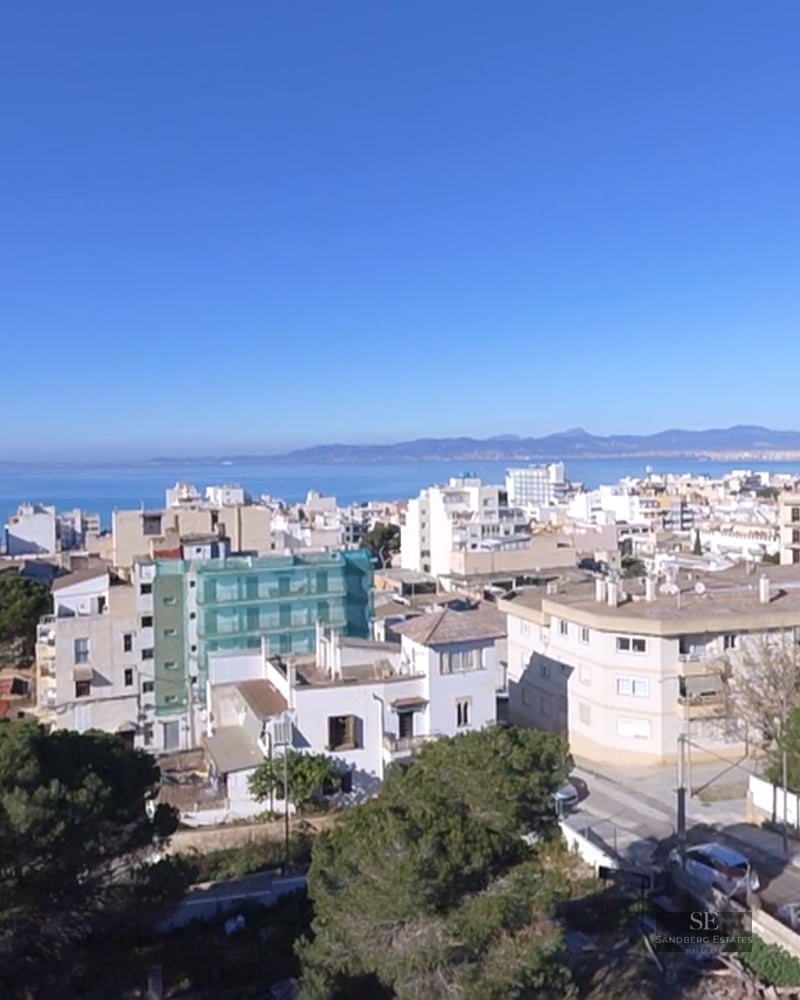 Elevated view of a Mediterranean coastal city with white buildings, green trees, and the blue sea on the horizon.