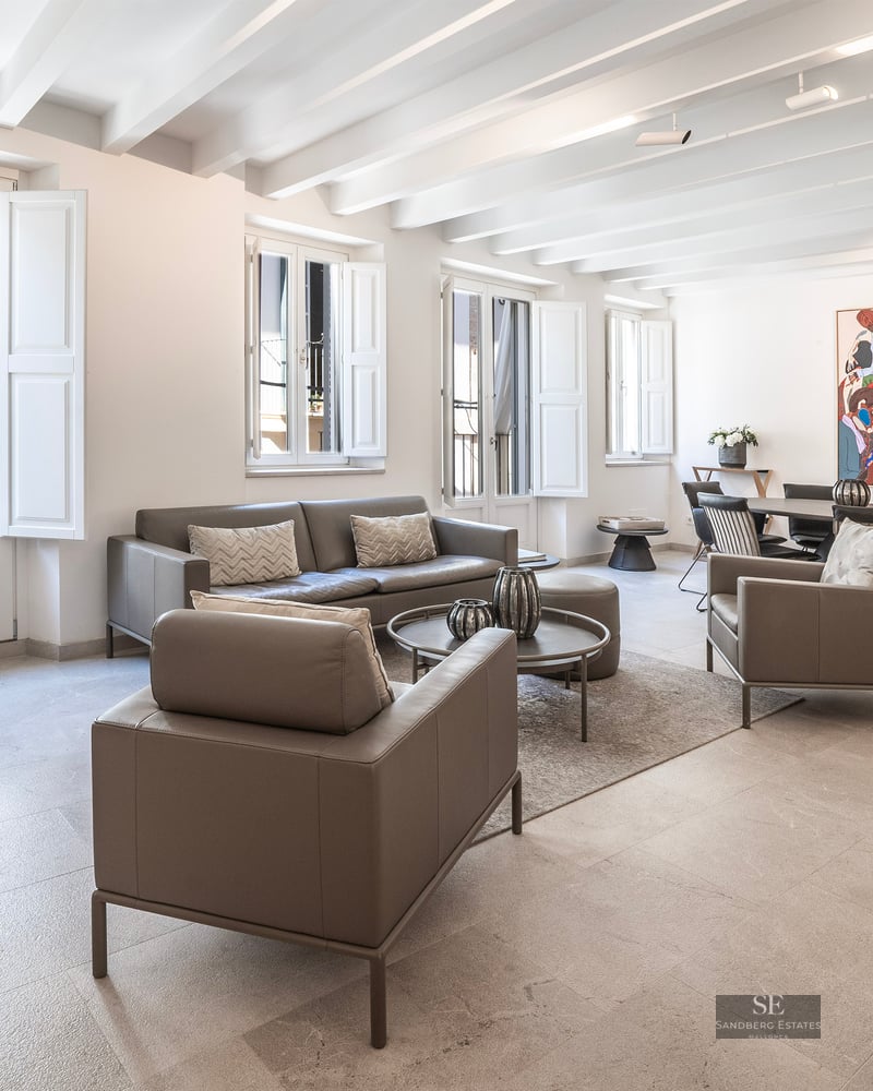 Salon lumineux avec canapés en cuir gris, plafond aux poutres blanches et coin repas avec une œuvre d'art colorée.