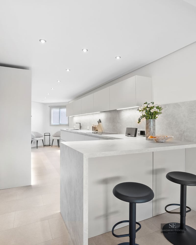 Une cuisine blanche moderne avec un comptoir en pierre, deux tabourets noirs et un éclairage LED intégré.