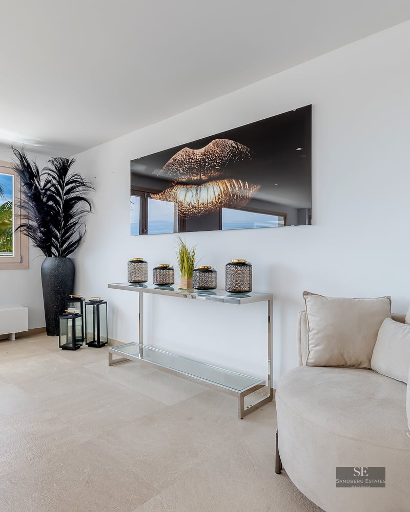 Modern lounge with a beige round armchair, gold lip wall art, and a view of palm trees through a window.