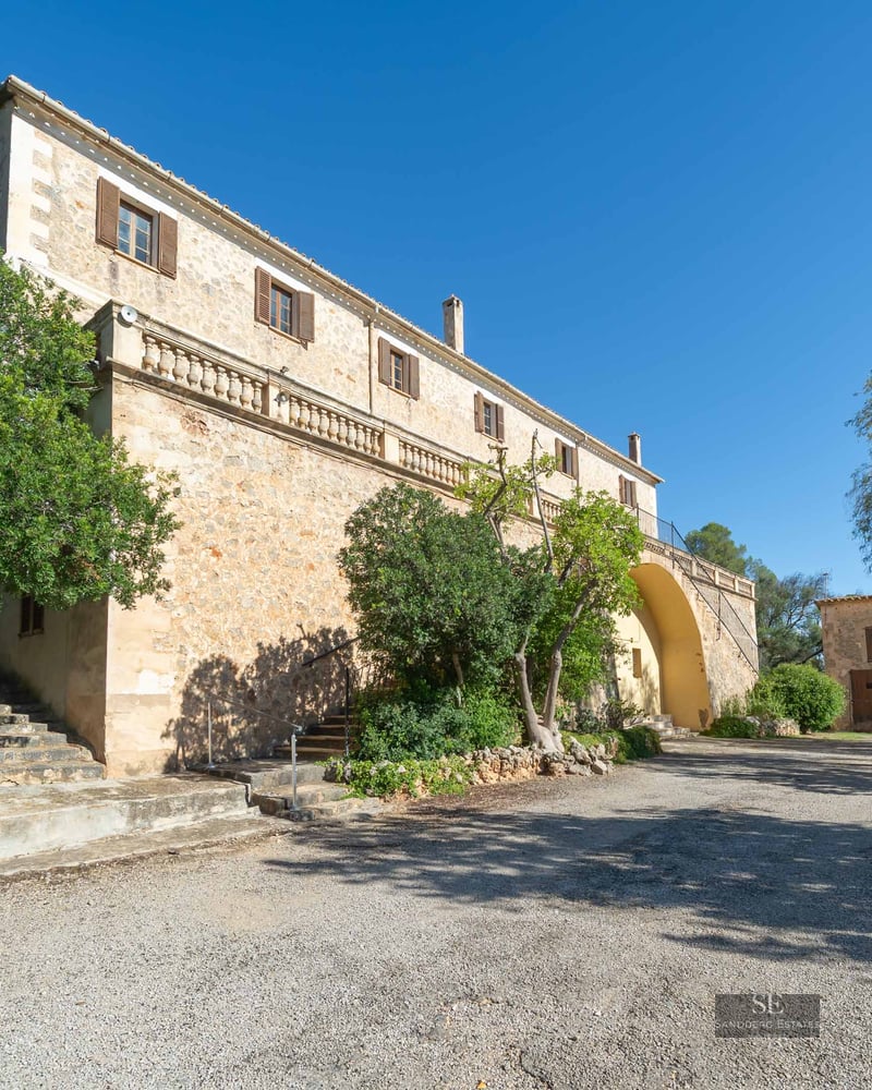 Grand stone Mediterranean villa with an elevated terrace, wooden shutters, and lush trees against a clear blue sky.