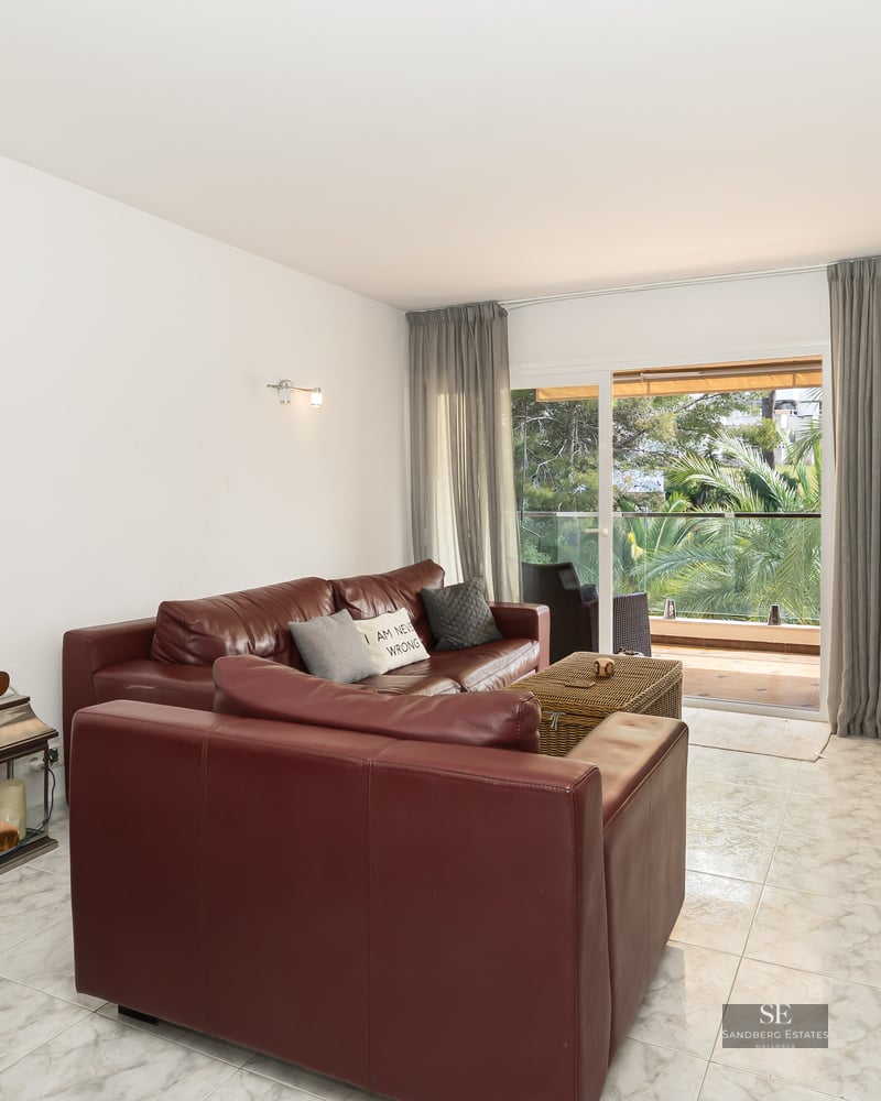 Modern living room featuring burgundy leather sofas, white marble floors, and a glass door view to a lush green balcony.