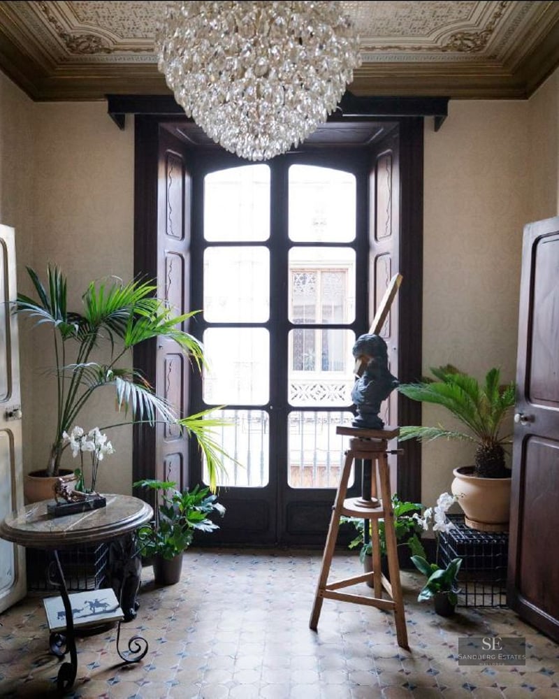Grand entrance with tall wooden doors, mosaic floors, a crystal chandelier, and indoor plants by a balcony window.