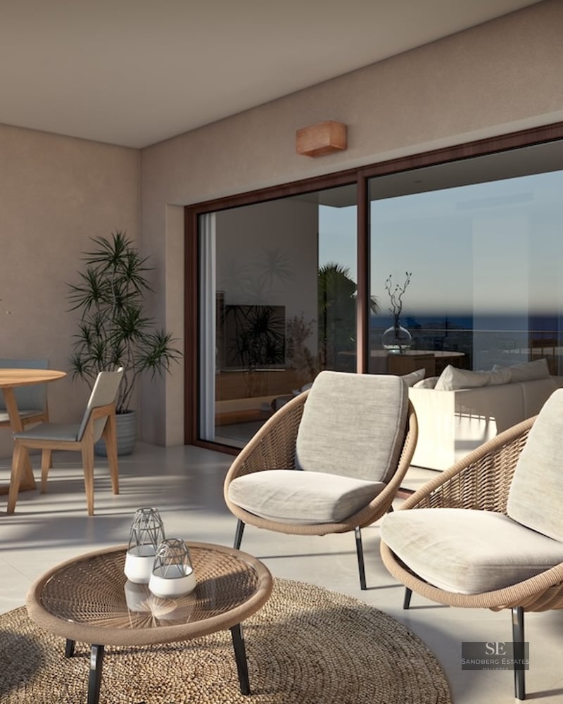Modern terrace with woven lounge chairs, wooden dining table, and large glass doors reflecting a sea view.