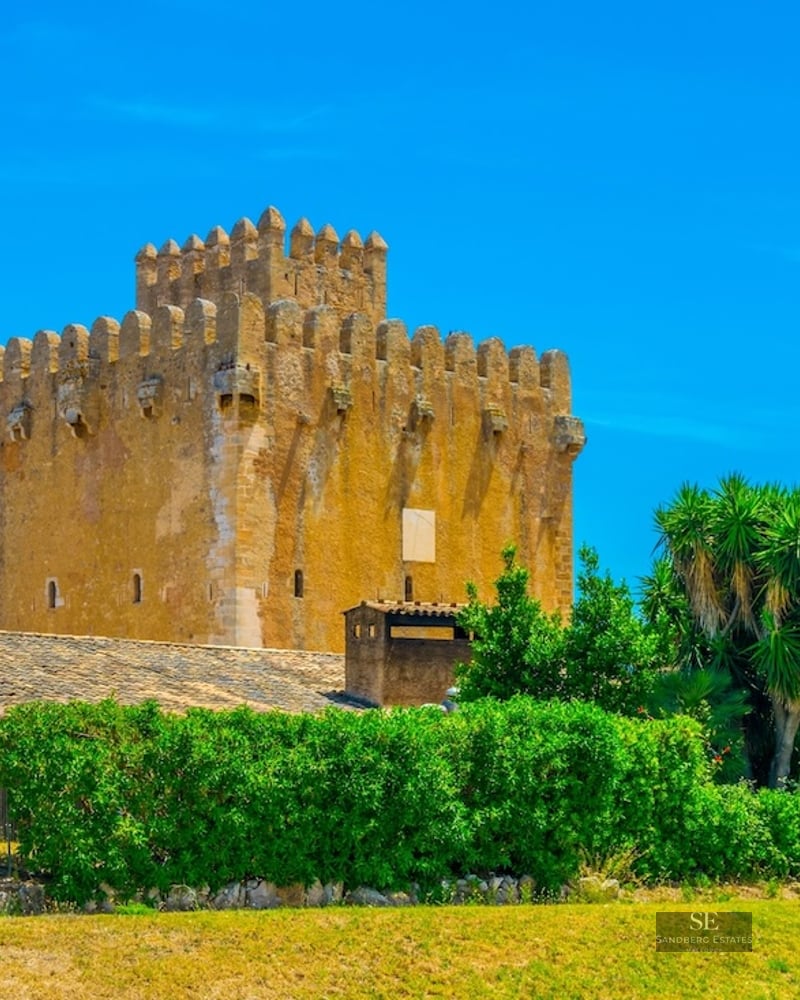 Golden stone medieval watchtower with battlements surrounded by lush greenery under a clear blue sky.