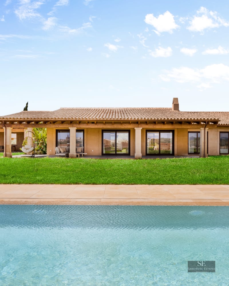 Large turquoise pool in front of a modern Mediterranean villa with a green lawn and terracotta roof under a blue sky.