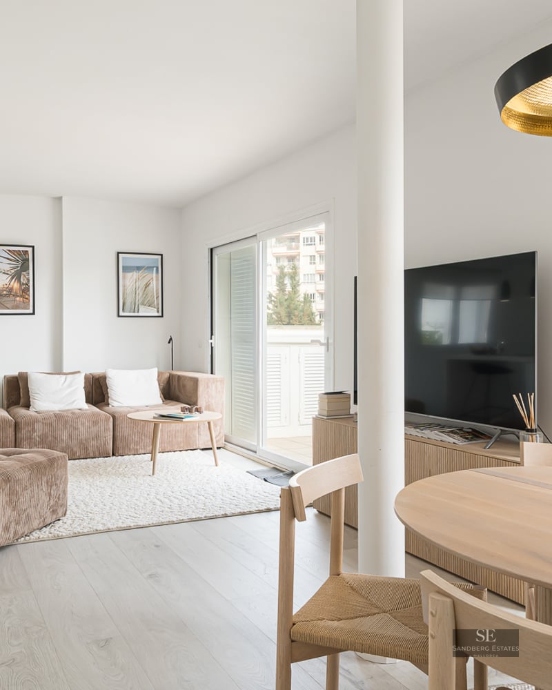 A sunlit living room featuring a brown modular sofa, wooden dining table, and large glass sliding doors leading to a balcony.