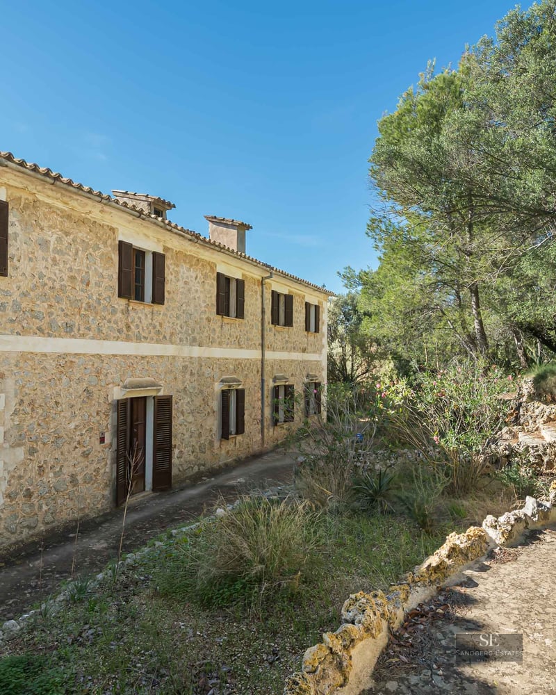 A two-story rustic stone building with dark brown shutters surrounded by lush green trees under a clear blue sky.