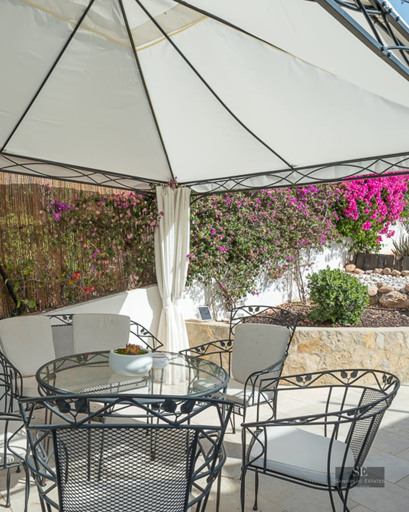 A white gazebo with a glass table and black wrought-iron chairs on a stone patio surrounded by pink flowers.