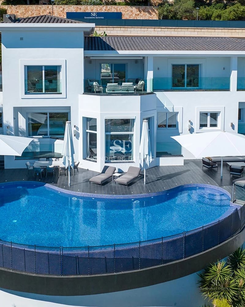 A multi-level white modern villa with a large curved infinity pool, expansive dark gray terrace, and white sun umbrellas.