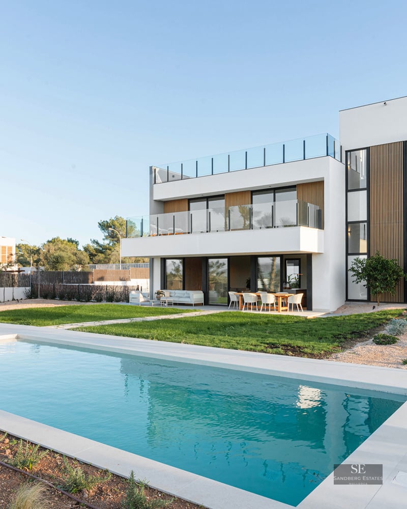 Modern white design villa featuring a rectangular swimming pool, manicured lawn, and spacious terraces under a clear sky.