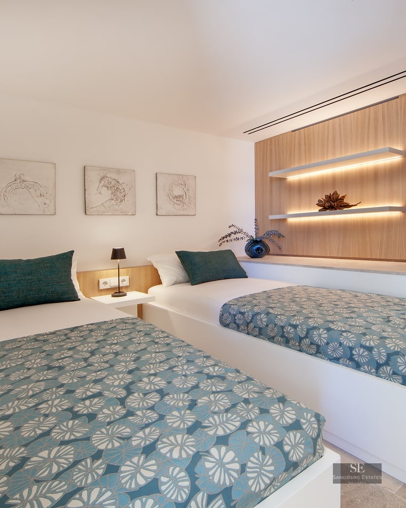 Modern bedroom featuring two single beds with teal patterned covers, wooden wall panels, and warm LED floating shelves.