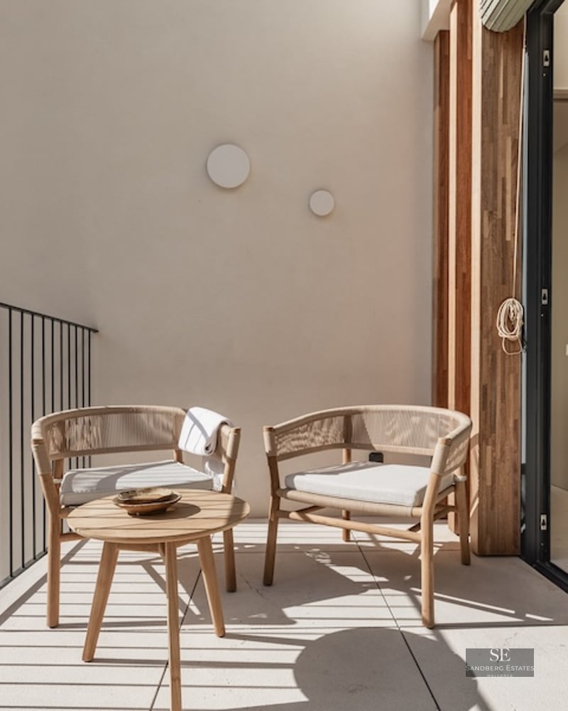 Sunlit balcony with designer woven chairs and a wooden coffee table overlooking a lush green forest.