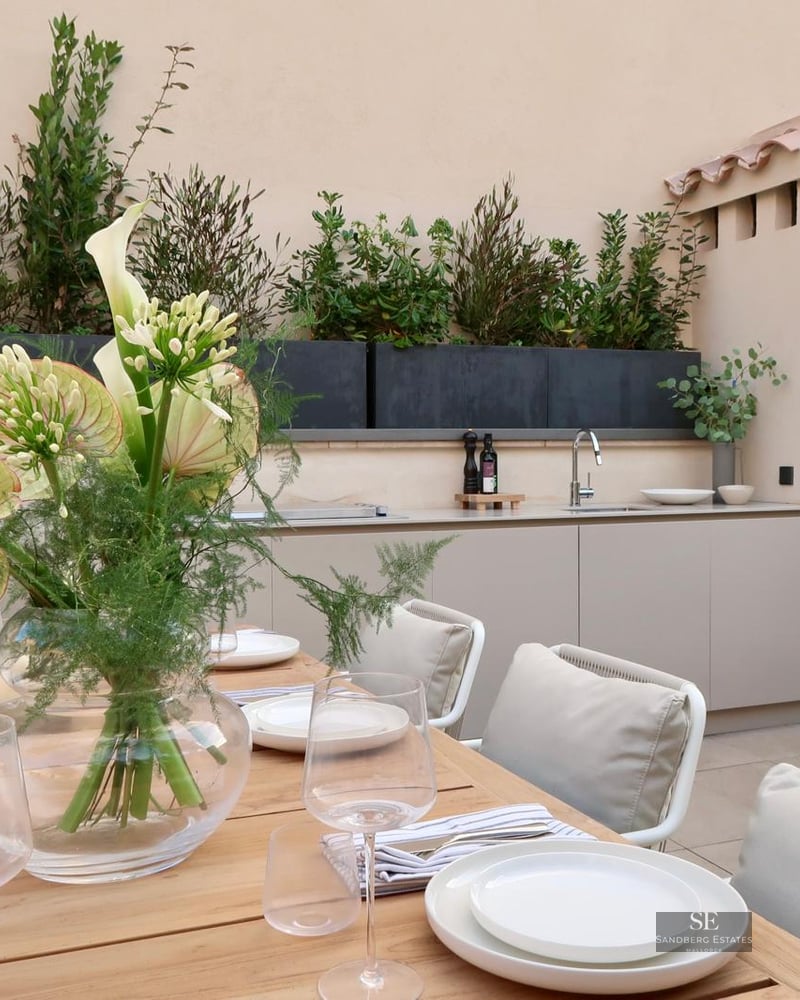 A wooden dining table set for a meal next to a modern outdoor kitchen and lush green planters.