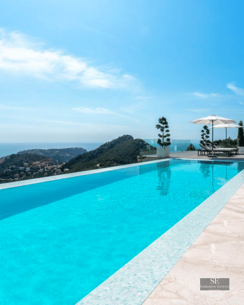 Turquoise infinity pool overlooking the sea and mountains, surrounded by light stone tiles and white umbrellas.