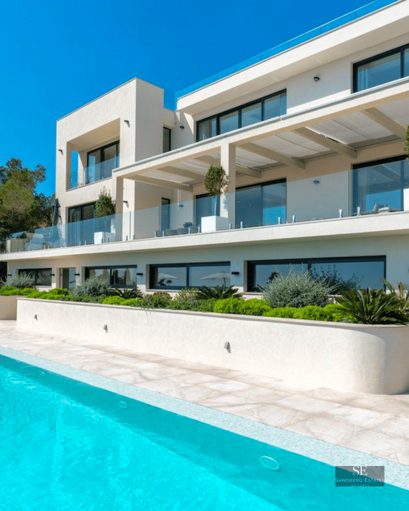 Large turquoise swimming pool in front of a modern white three-story villa with glass balconies and stone stairs.
