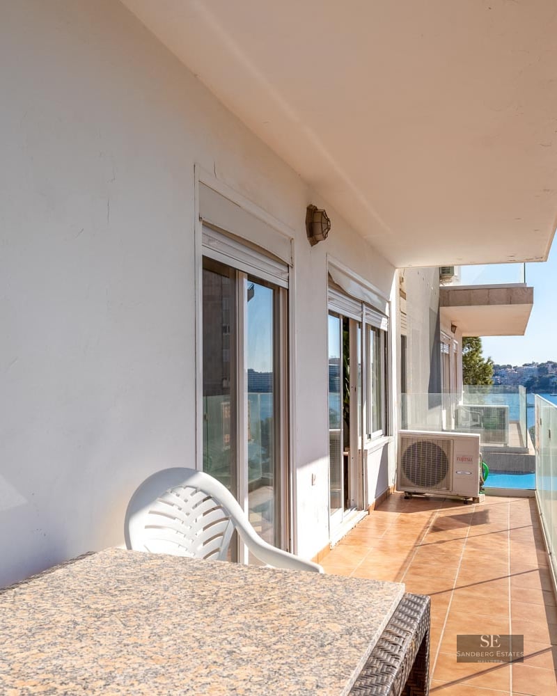 Balcony with glass railing and stone table overlooking a blue bay with boats and coastal buildings.
