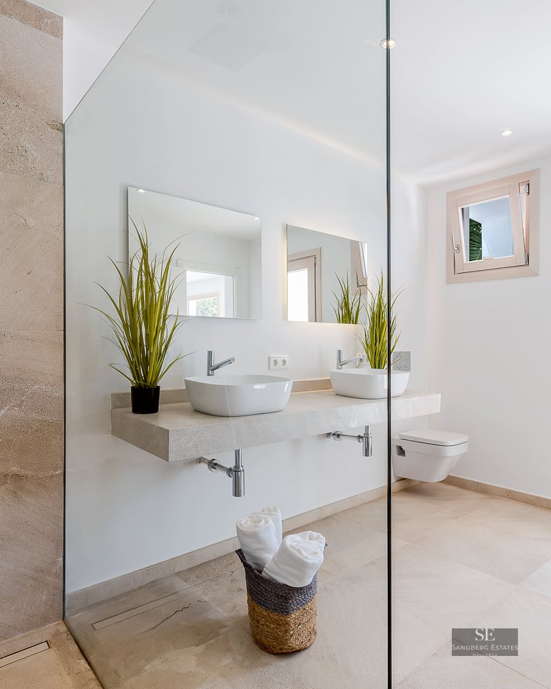 Bright modern bathroom with stone tiles, glass shower, double vessel sinks on a floating vanity, and garden view.
