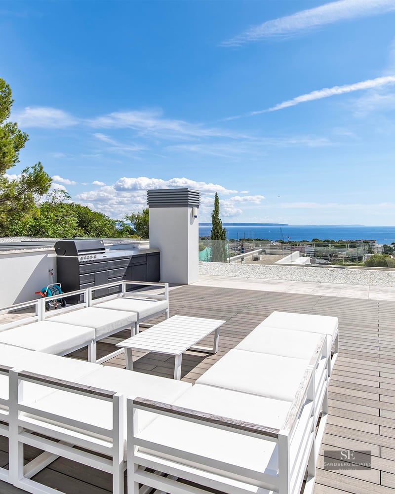 Spacious modern rooftop terrace with white lounge seating, glass railings, and a wide view of the sea under a blue sky.