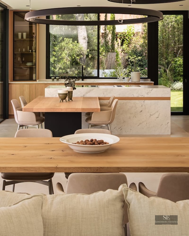 Modern kitchen with a large marble island, integrated wooden dining tables, and floor-to-ceiling windows showing a garden.