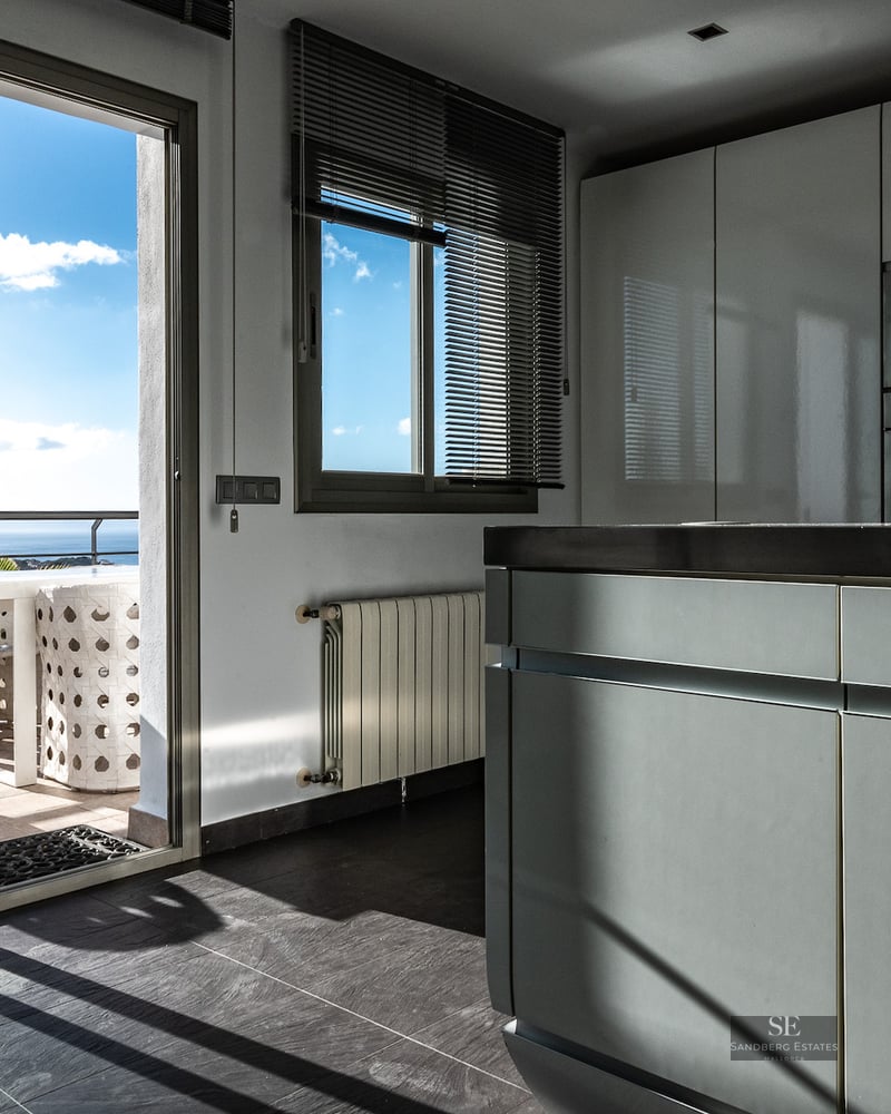 Minimalist white kitchen with an island and large sliding glass doors opening to a sunny terrace with sea views.