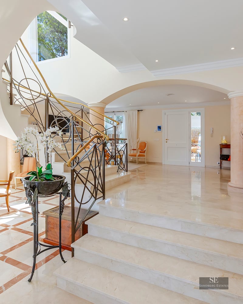 A bright luxury entrance hall featuring a white marble staircase with ornate iron railings and arched architecture.