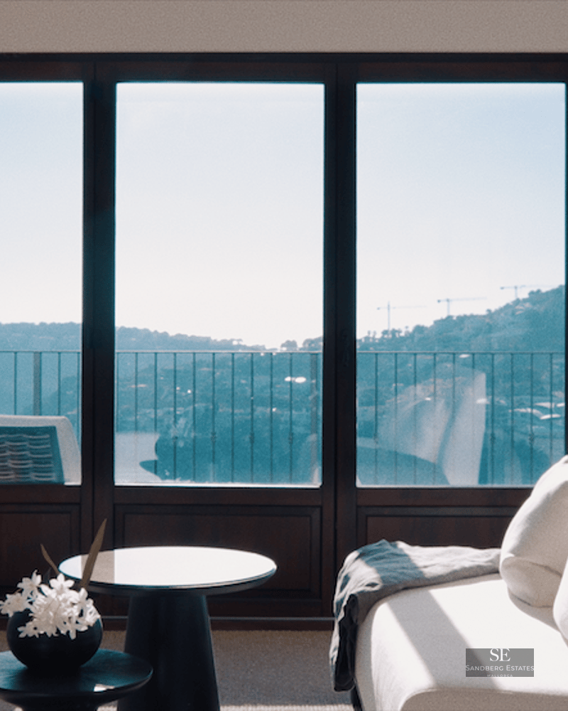 Modern master bedroom featuring a white sofa, canopy bed, and floor-to-ceiling windows overlooking the sea and hills.