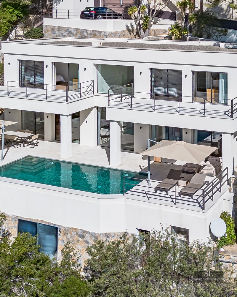 Elevated view of a white multi-level modern villa with a turquoise swimming pool on a stone terrace.