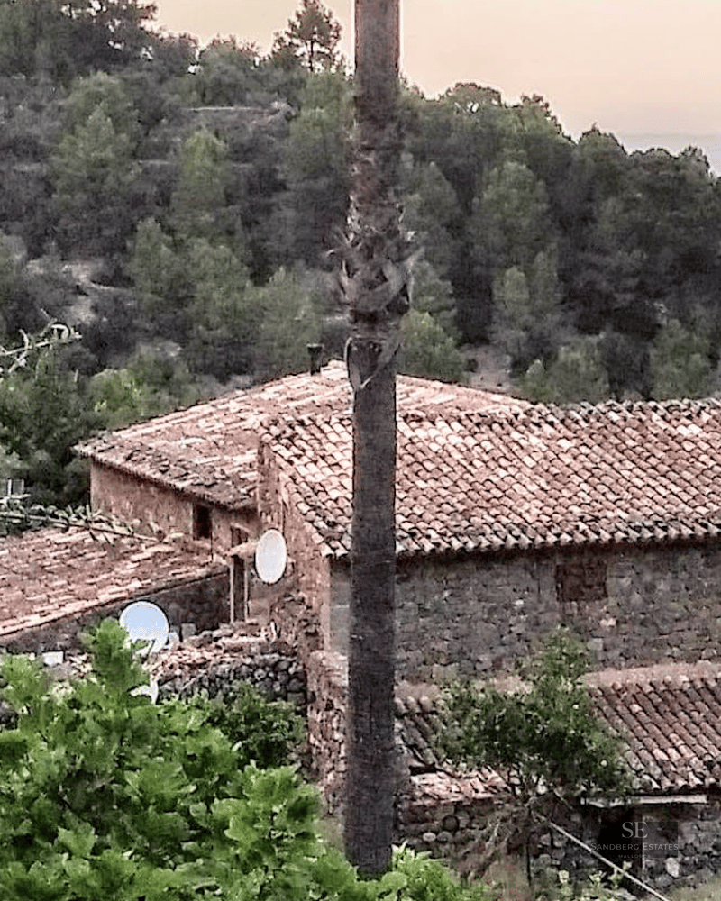 Traditional stone building with terracotta tiled roofs nestled in a lush green hillside overlooking the sea.