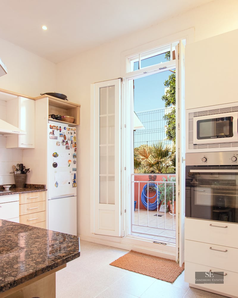 Modern white kitchen featuring brown granite countertops and an open door leading to a sunny balcony.