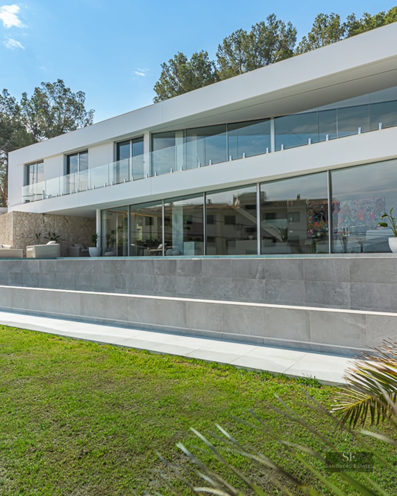 White modern villa with large windows, infinity pool, and green lawn under a sunny sky.