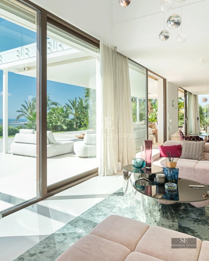 Spacious living room with floor-to-ceiling windows overlooking the sea. Modern design, light wood floors, and minimalist furniture. Abundant natural light.