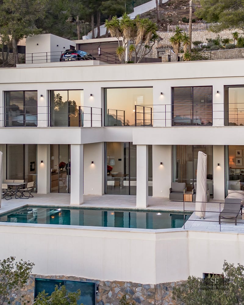 Modern white two-story villa on a hillside with a rectangular pool and multiple terraces at dusk.