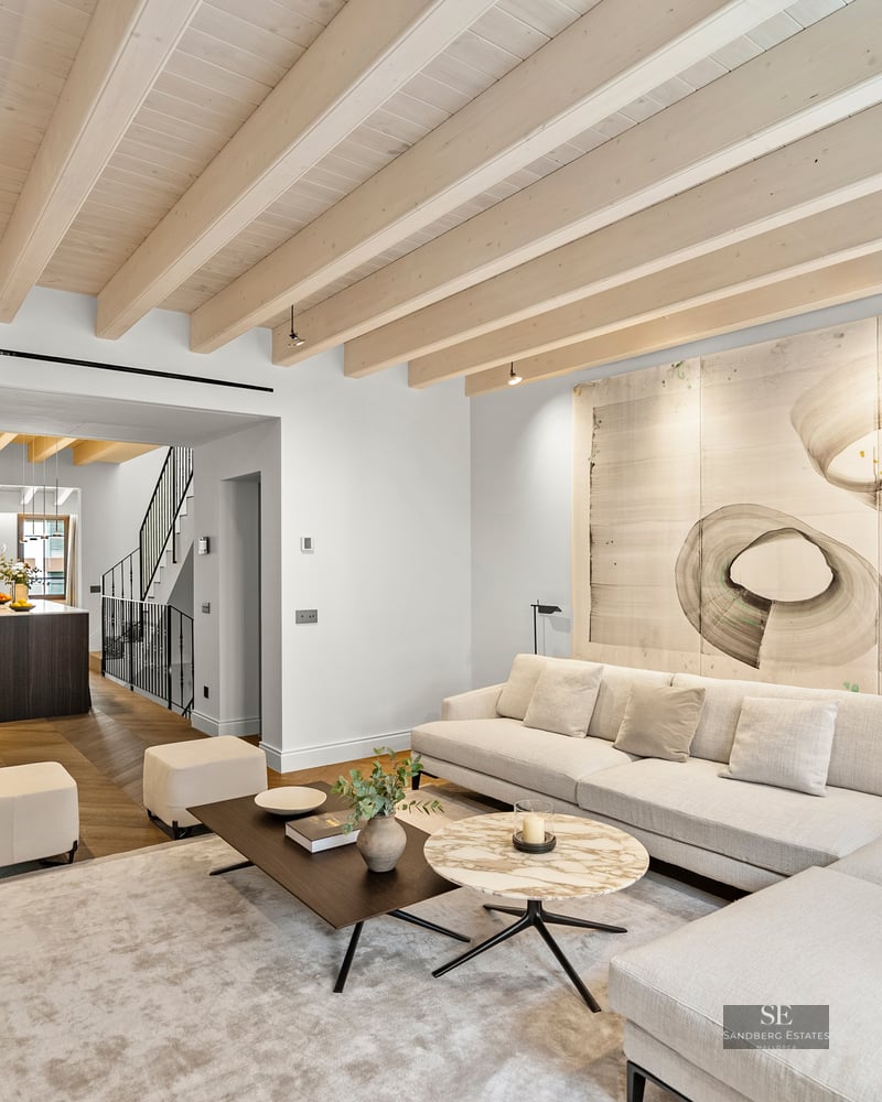 A bright living room with a large white sectional sofa, wooden ceiling beams, and abstract art above the couch.