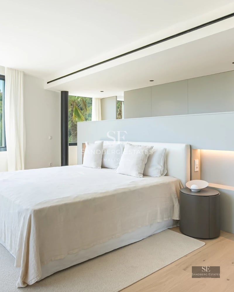 Spacious living room with floor-to-ceiling windows, light wood floors, and a fireplace. Minimalist furniture and abundant natural light. Garden view.