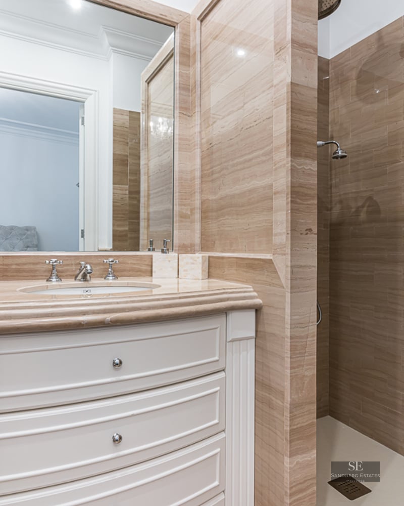 Luxury bathroom featuring beige travertine tile walls, a white curved vanity with marble top, and a walk-in shower.